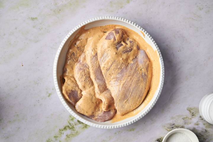 Raw chicken breasts in a creamy orange marinade are in a white, ridged dish on a light marble surface.
